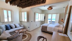A spacious room with a sofa, a bed, a wooden table, and a chair. There are windows with curtains, a ceiling fan, and a hanging light fixture. - LIFE SUITES (Mykonos)