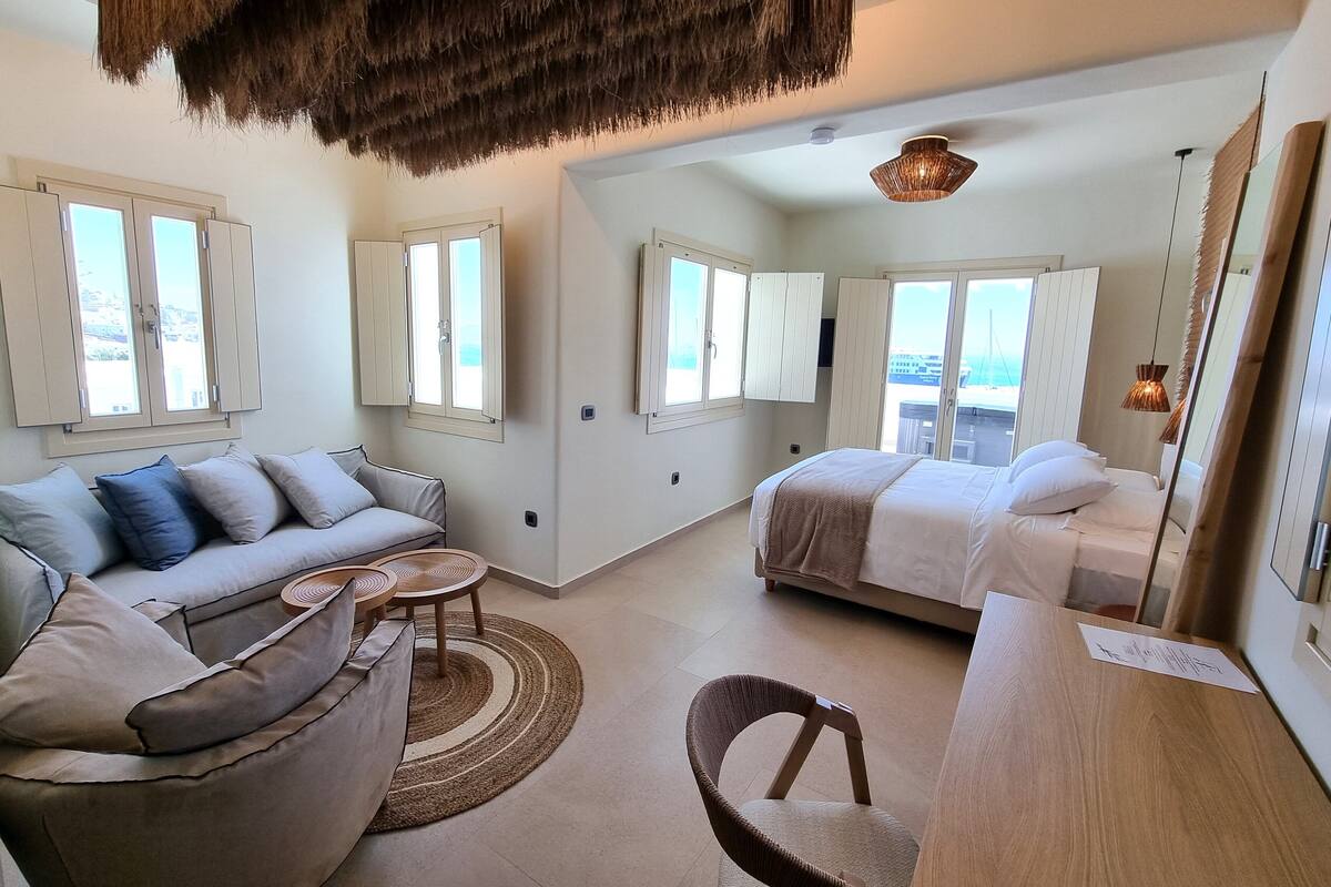 A spacious room with a sofa, a bed, a wooden table, and a chair. There are windows with curtains, a ceiling fan, and a hanging light fixture.