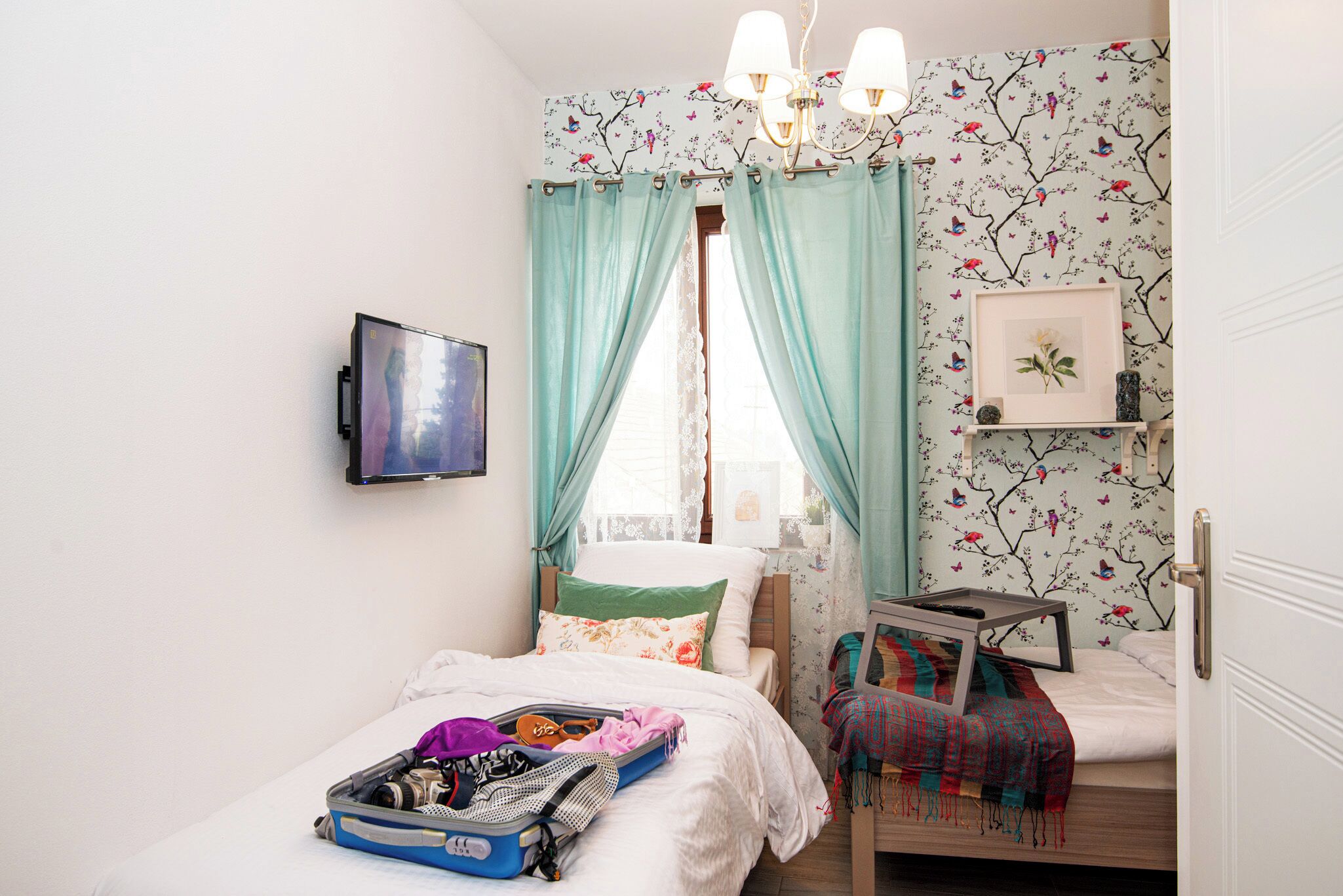 A bedroom with a bed, a suitcase, a TV, a window with curtains, a small table, and a wall with floral wallpaper.