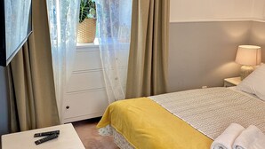 Ein Schlafzimmer mit einem Bett, einem Nachttisch, einem Fernseher, einem kleinen Tisch und einem Fenster mit Vorhängen.
