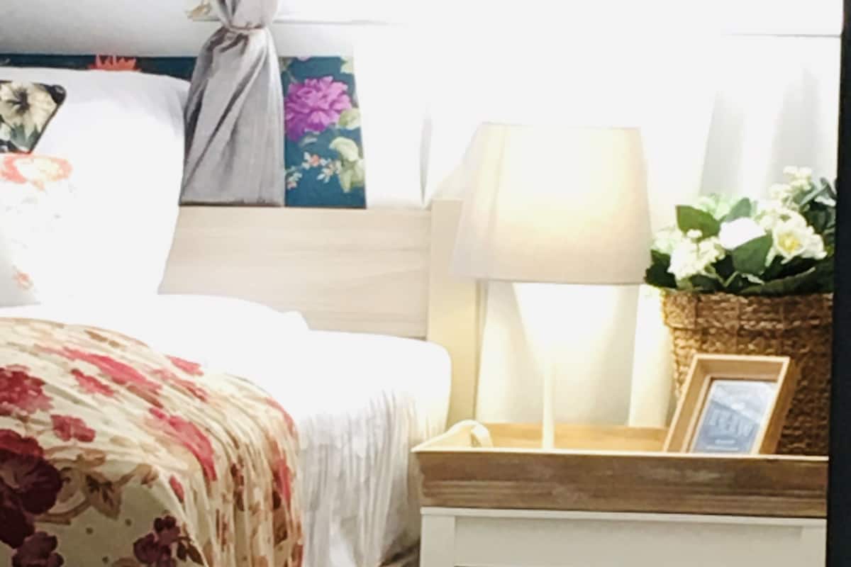 Een netjes opgemaakt bed met een bloemenbedekking, een nachtkastje met een lamp, een ingelijst beeld en een klein plankje met een mand en een bloevas.
