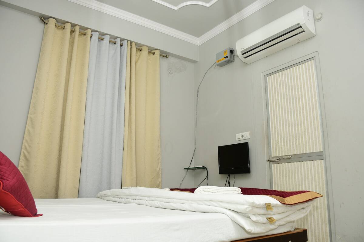 A neatly made bed with white bedding, a red pillow, and a beige curtain.
