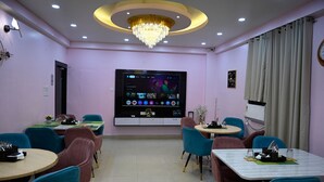 A hotel dining area with multiple tables and chairs, a large flat-screen TV on the wall, and a chandelier. - Hotel Buddha International (Patna)