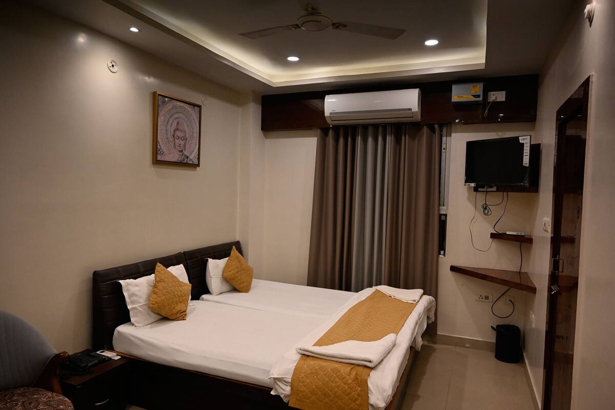 A hotel room with a bed, a ceiling fan, an air conditioning unit, a television, and a small trash bin.