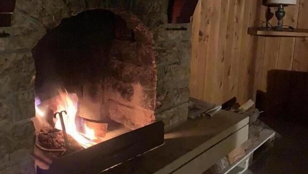 A stone fireplace with a fire burning inside, wooden beams above, and a shelf with a lamp to the right. - 2-bedroom apartment (Bosco Chiesanuova)