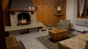 A cozy living room with a stone fireplace, wooden walls, a patterned armchair, and a wooden coffee table. - 2-bedroom apartment (Bosco Chiesanuova)