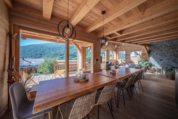 A spacious wooden dining area with a long table and wicker chairs, large windows offering a view of a forested mountain, and a stone wall on one side.