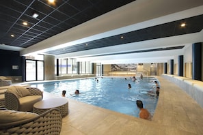 An indoor swimming pool with multiple lanes, lounge chairs, and a view of mountains through large windows.