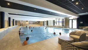 Une piscine intérieure dotée de plusieurs couloirs, d’un espace détente avec des fauteuils confortables et d’une vue sur les montagnes grâce à de grandes fenêtres.