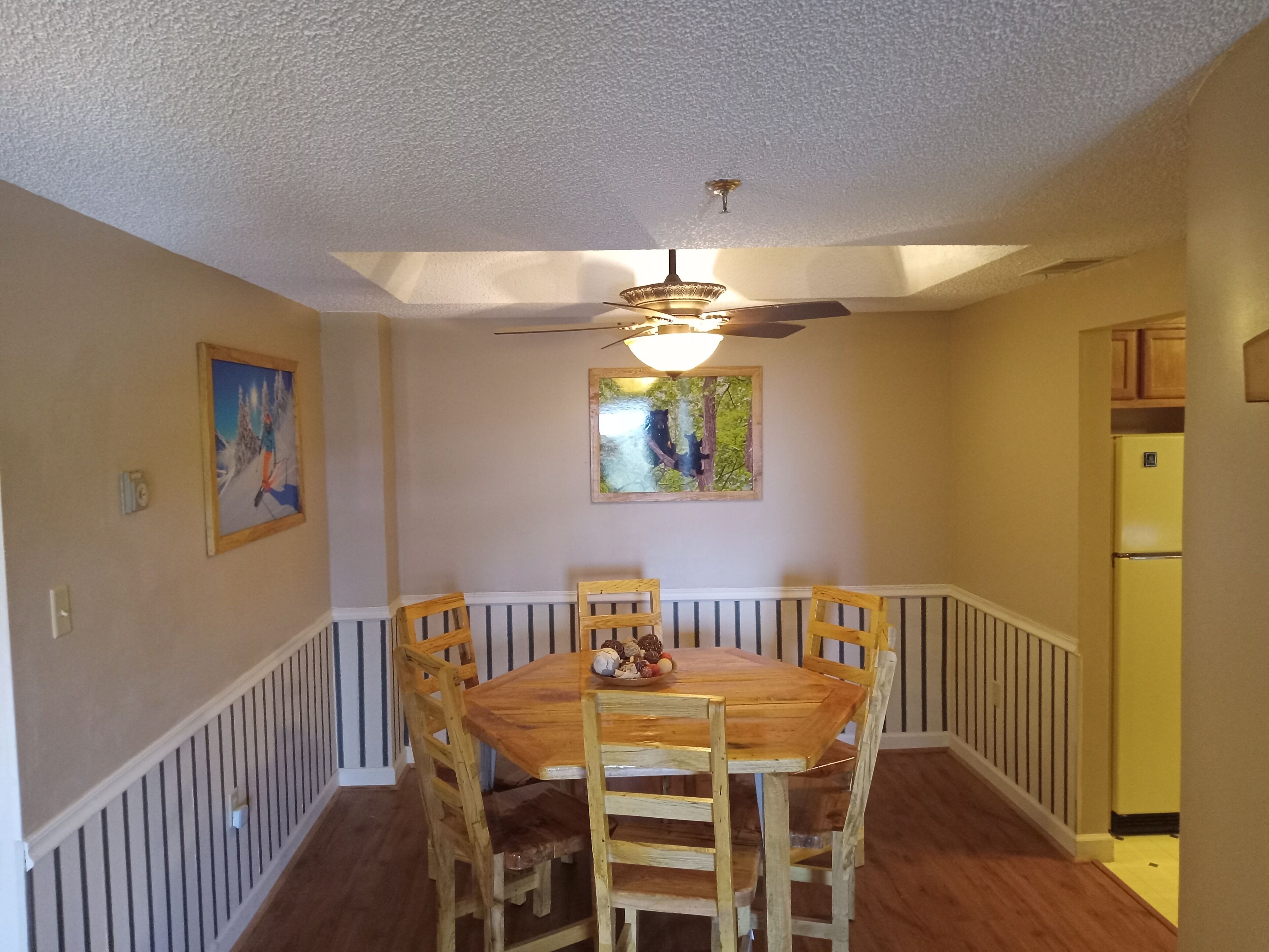 A dining area with a wooden table and chairs, a ceiling fan, and two wall-mounted paintings.