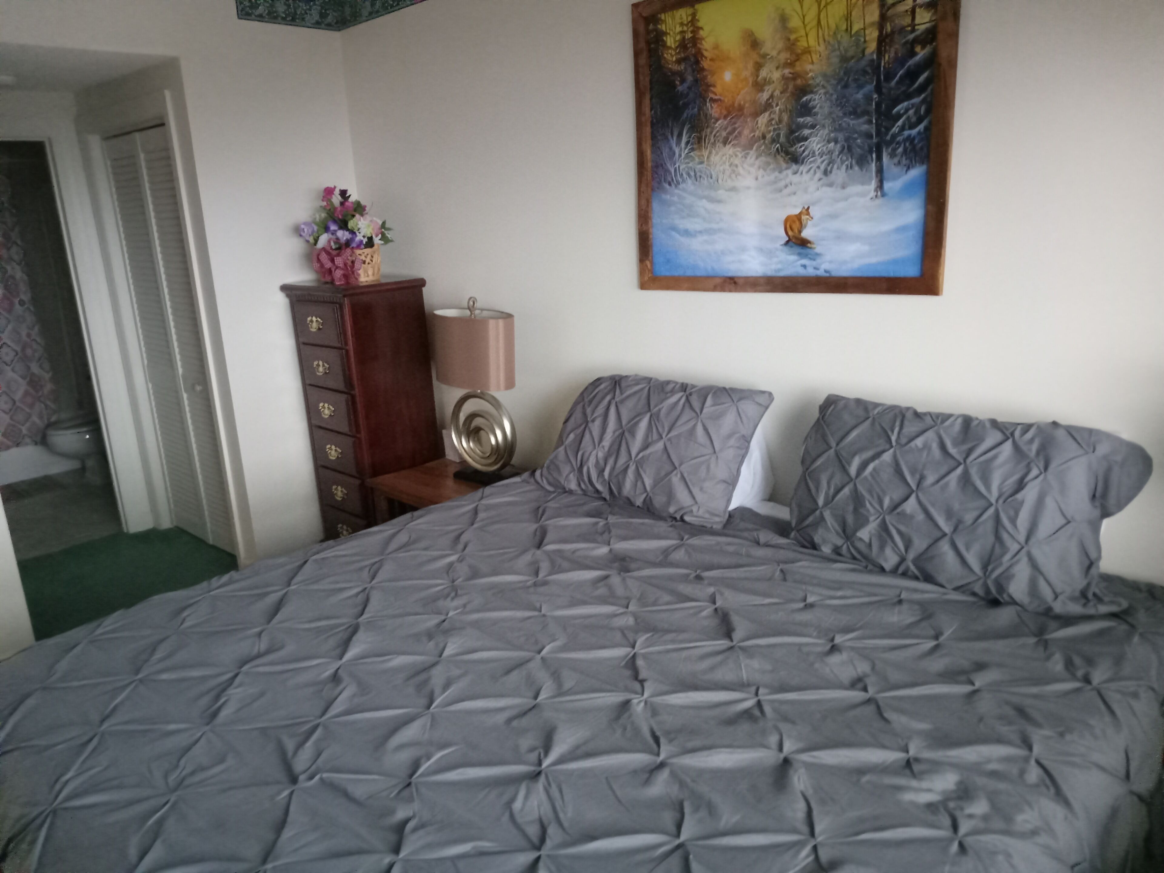 A bedroom with a large bed, a dresser, a lamp, and a painting of a fox in a snowy forest.