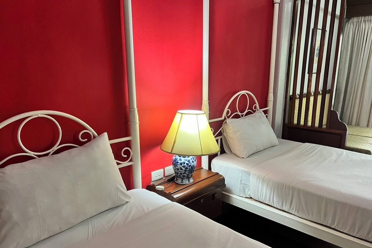 A hotel room with two single beds, a bedside table with a lamp, and a window with vertical blinds.