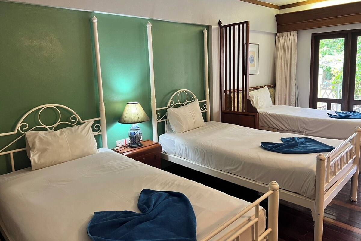 A hotel room with two single beds, a nightstand with a lamp, a wooden headboard, and a window with curtains.