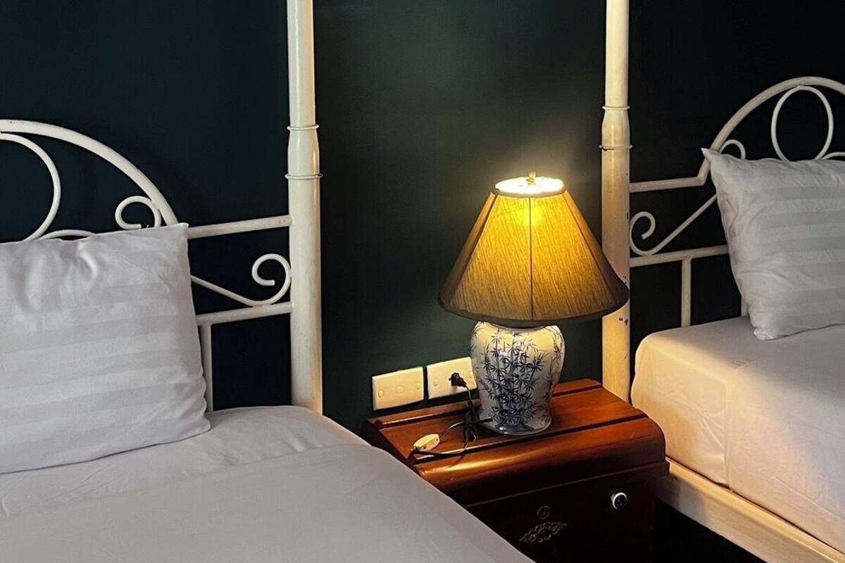 A room with two beds, each with white headboards and pillows, a bedside table with a lamp, and framed pictures on the wall.