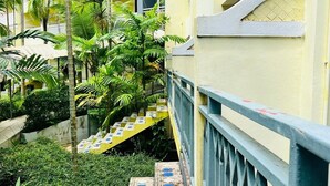 Una escalera con peldaños amarillos y barandillas azules, rodeada de abundante vegetación y un edificio amarillo con balcones.