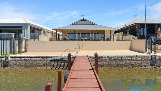 A waterfront property with a wooden dock leading to a covered area with a pool, surrounded by a stone wall and a fence.