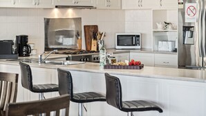 A modern kitchen with white cabinets, a stainless steel refrigerator, a microwave, and a gas stove. There are two black bar stools and a wooden cutting board with utensils.