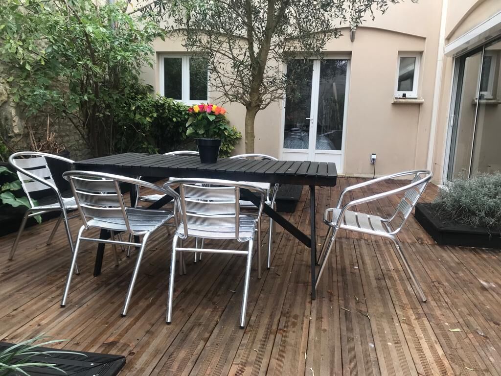 Een buitenpatio met een houten terras, een zwarte tafel en witte metalen stoelen.