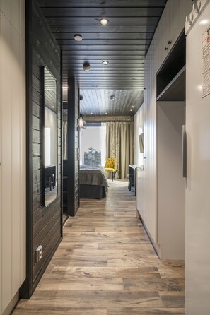 A narrow hallway with wooden flooring, a bed with a yellow chair, and a large mirror on the left.