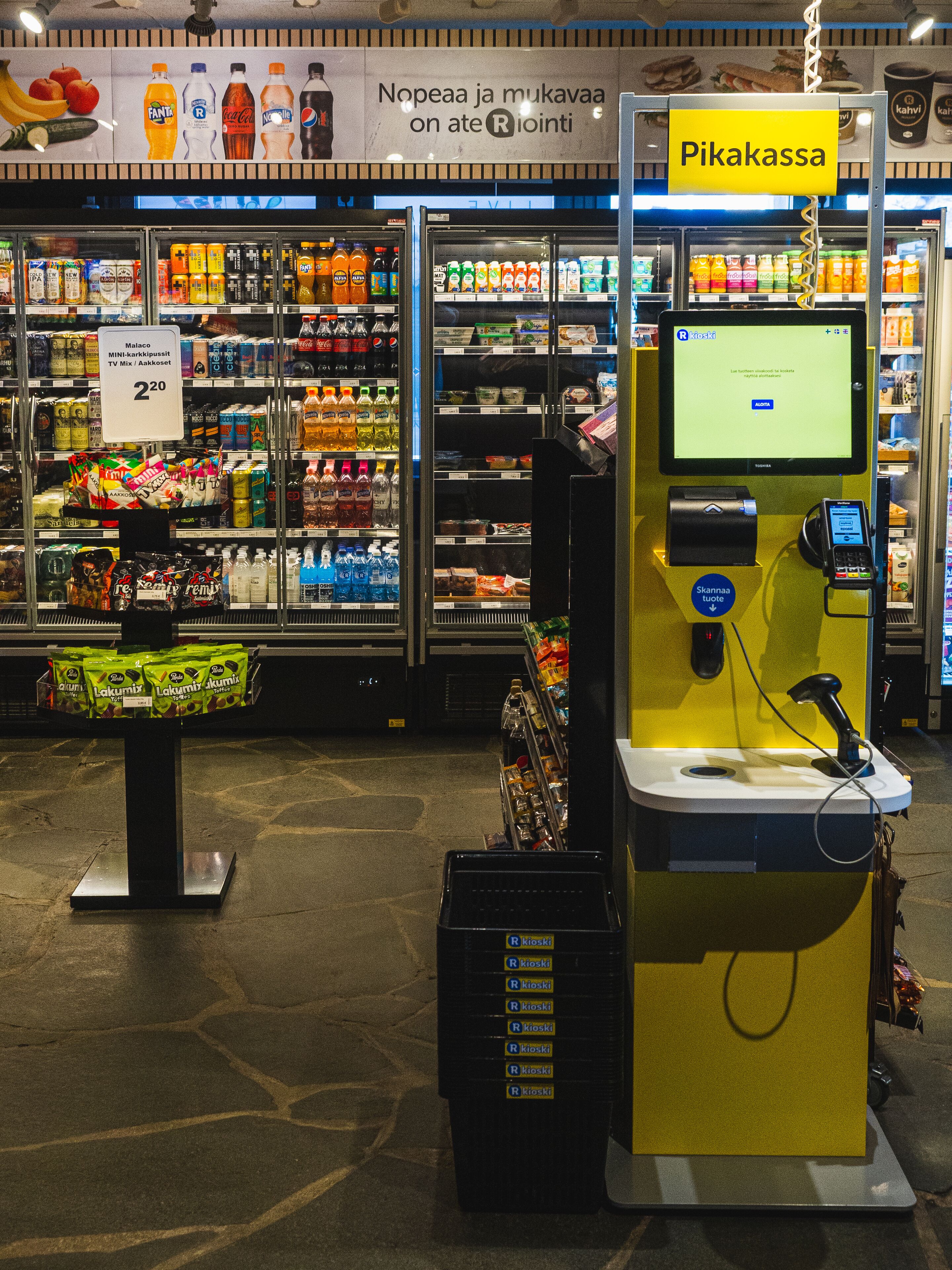 A self-service beverage kiosk with a payment terminal and a digital screen, surrounded by refrigerated beverage displays and a variety of snacks.
