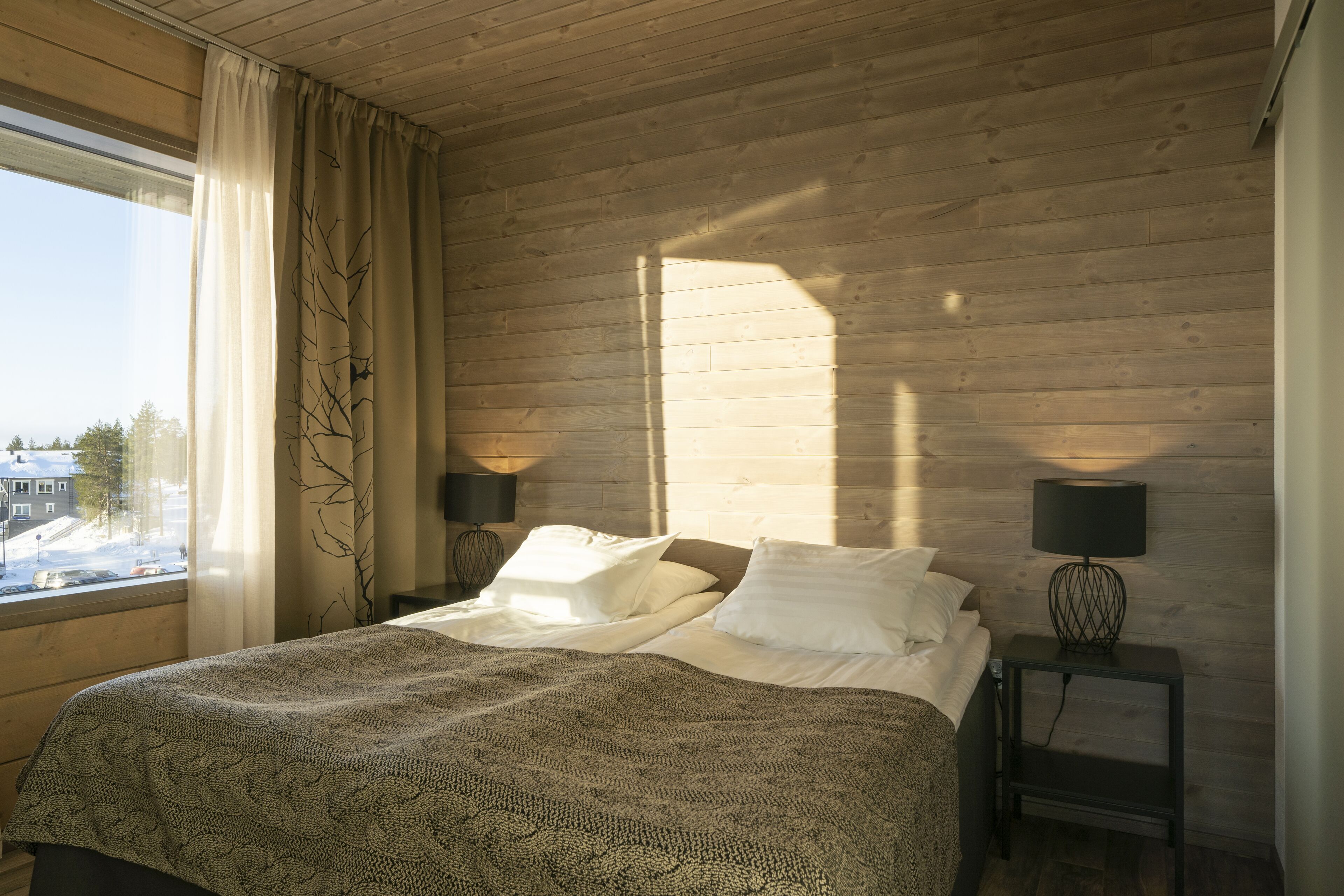 Ein Schlafzimmer mit Holzboden, einem großen Fenster mit Vorhängen, einem Bett mit weißen Leinen und zwei Nachttischen mit Lampen.