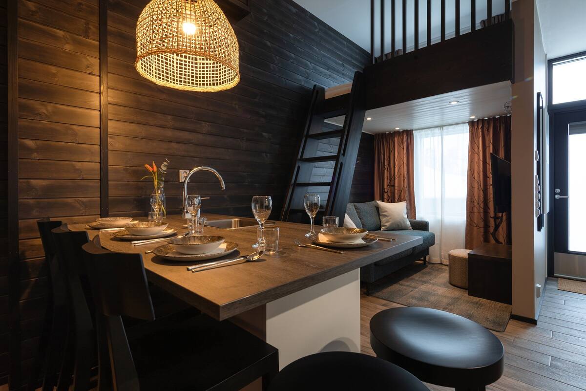 A modern dining area with a wooden table set for four, a hanging light fixture, and a view of the living area with a sofa and curtains.