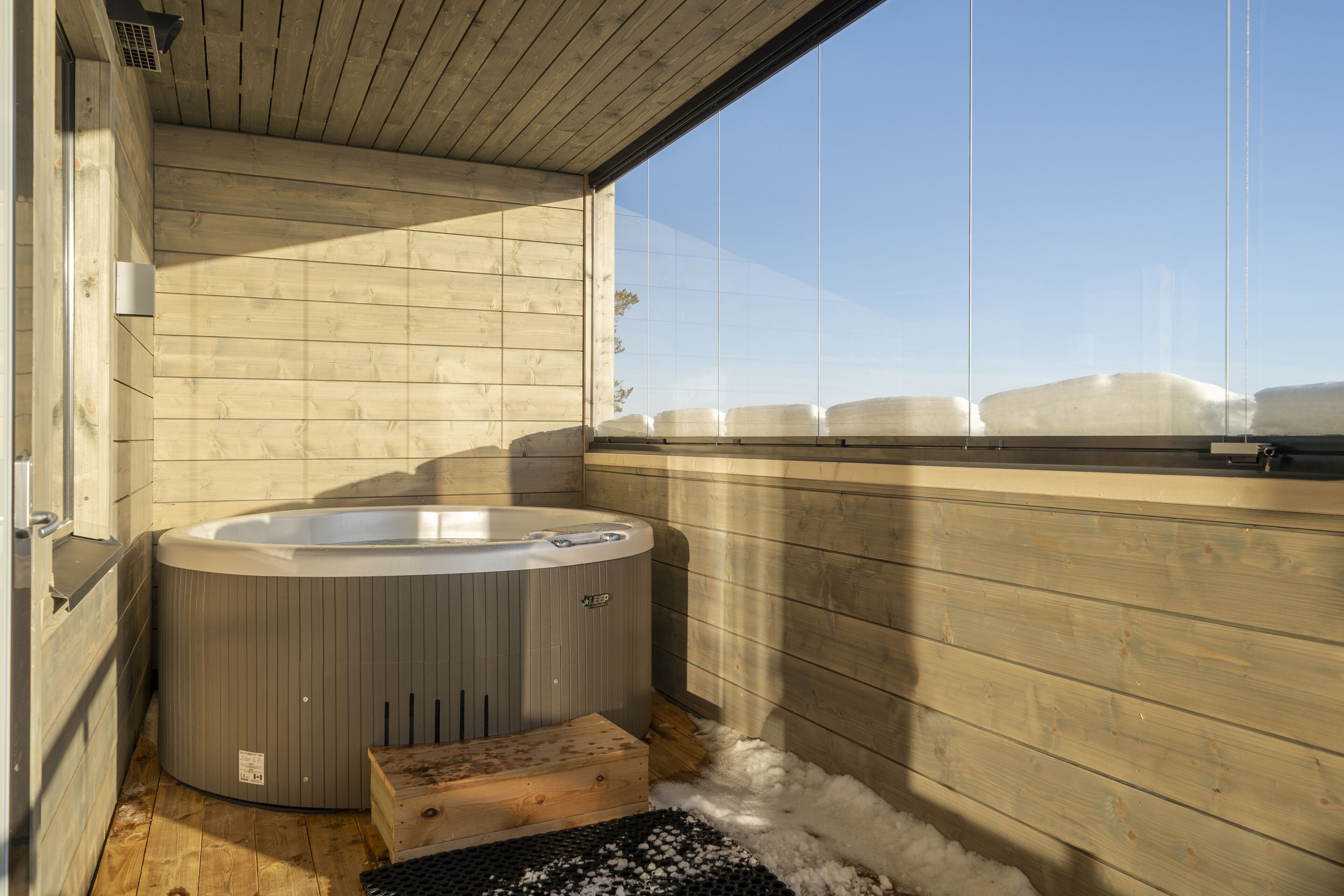 A wooden cabin with a hot tub, wooden flooring, and a window overlooking a snowy landscape.