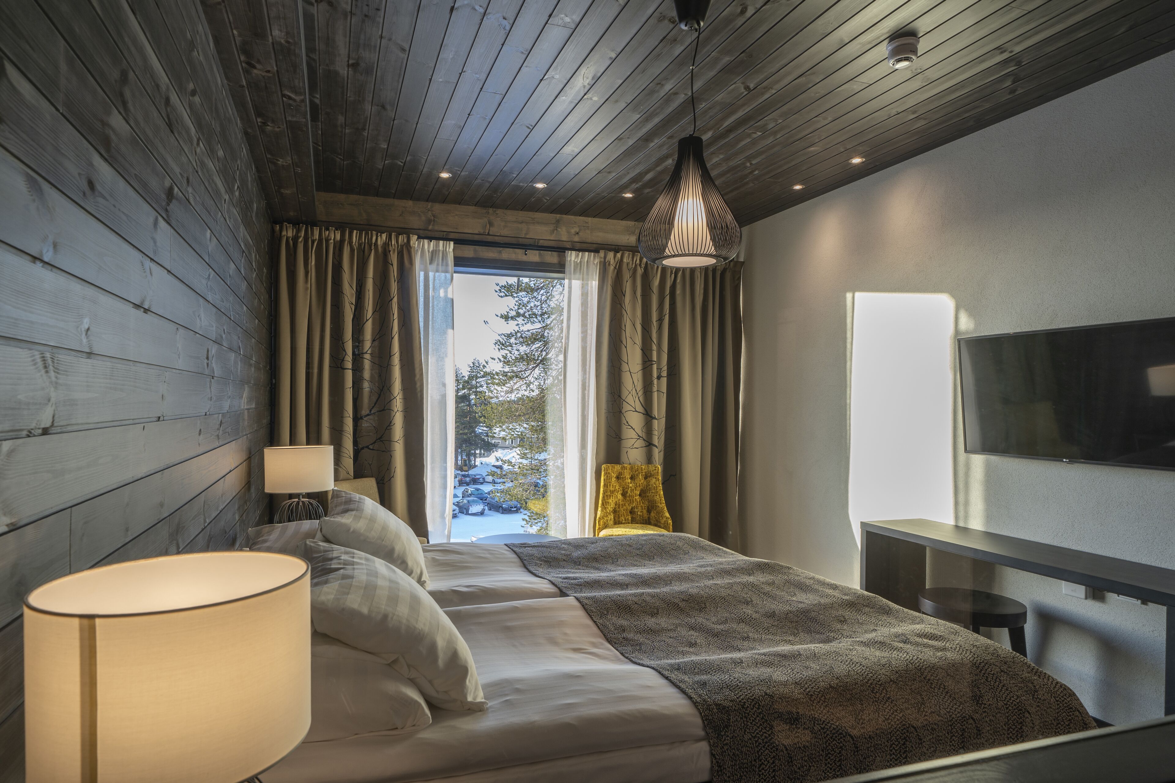 A modern bedroom with a wooden ceiling, a large bed with a blanket, a bedside lamp, and a TV on the wall.