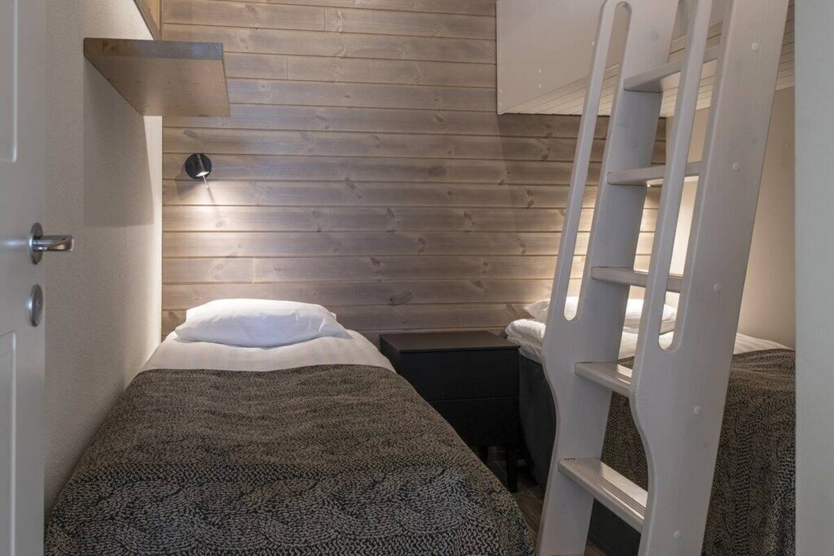 A compact room with a bunk bed, a bedside table, a lamp, and a wooden ceiling.