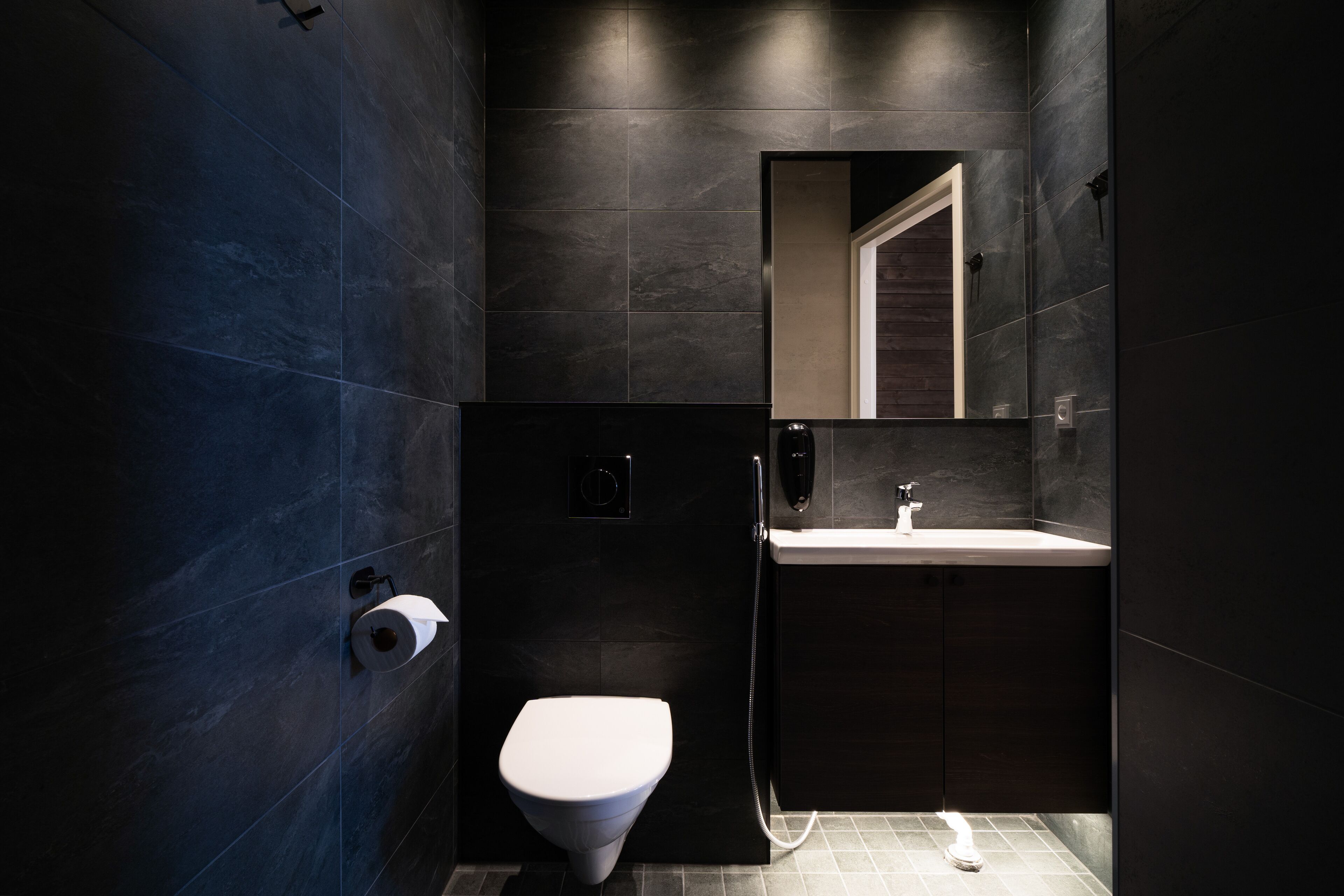 Een moderne badkamer met donkere tegels, een toilet, een wastafel en een spiegel.