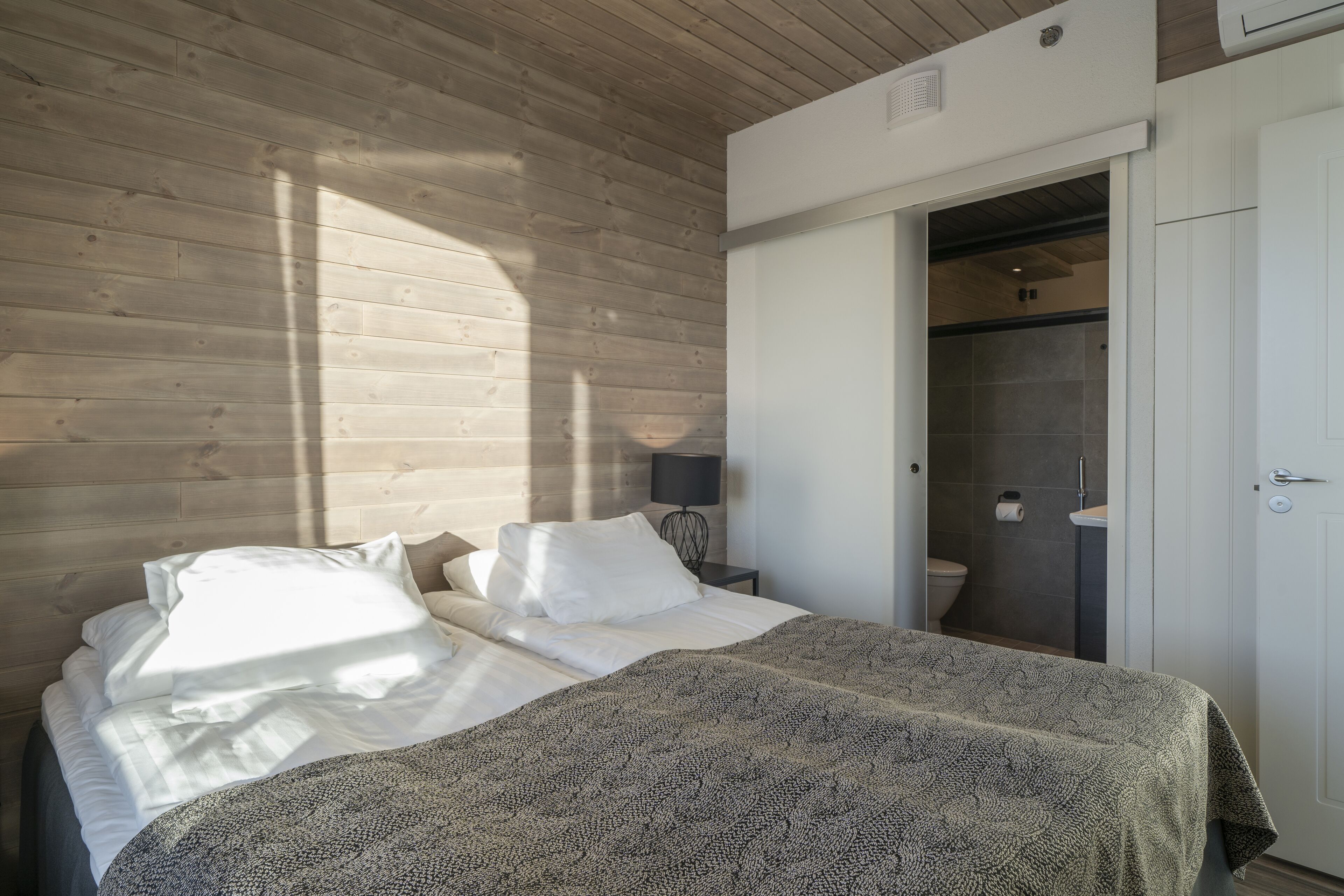 Un dormitorio con pared de madera, una cama con ropa blanca, una manta estampada, una mesita de noche con lámpara y una puerta abierta que da a un baño.