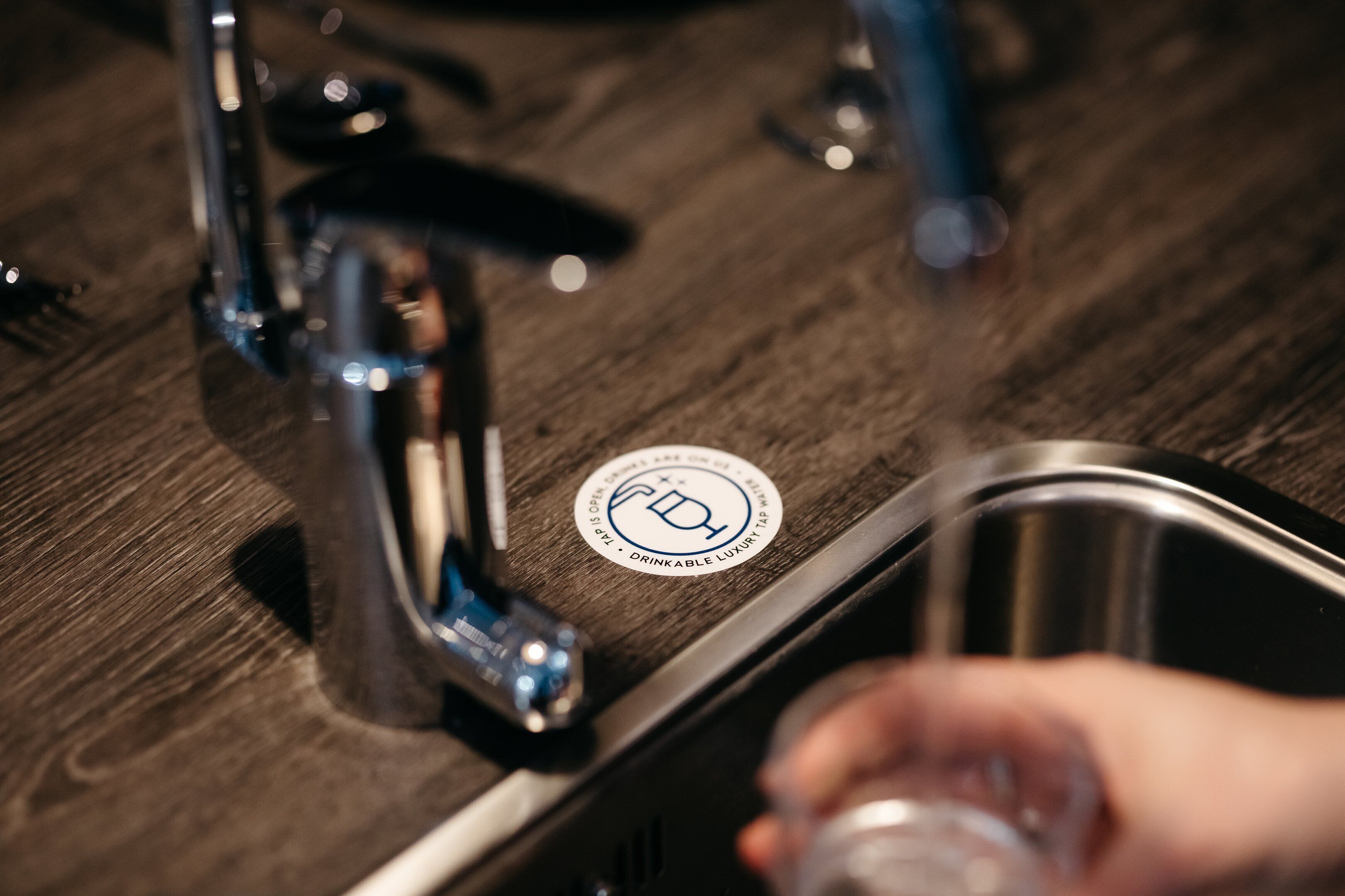 Primer plano de un lavamanos de cocina, una mano sosteniendo un vaso y un logotipo circular en la encimera.