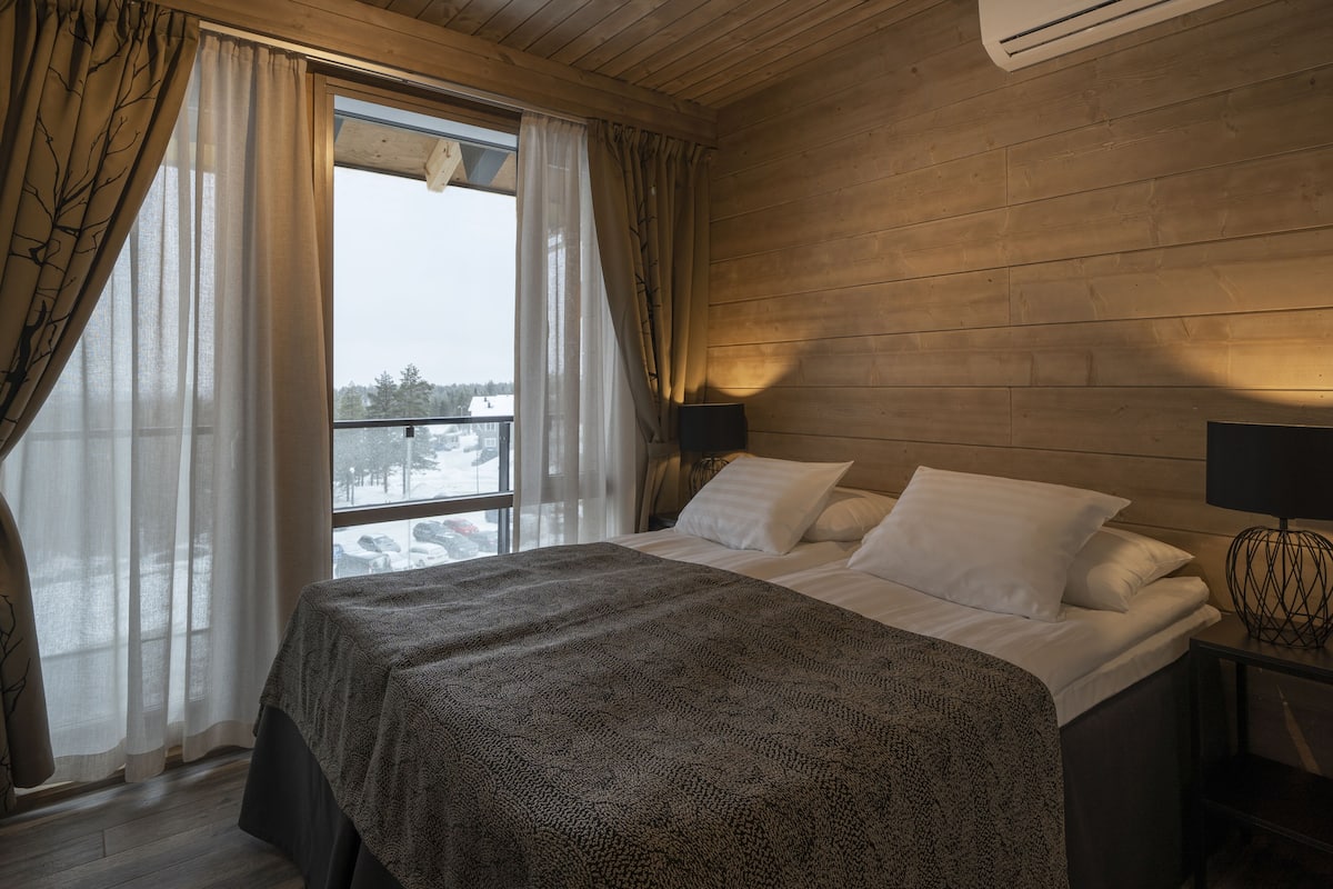 A bedroom with a bed, white pillows, a dark blanket, a window with sheer curtains, and a wall-mounted air conditioner.