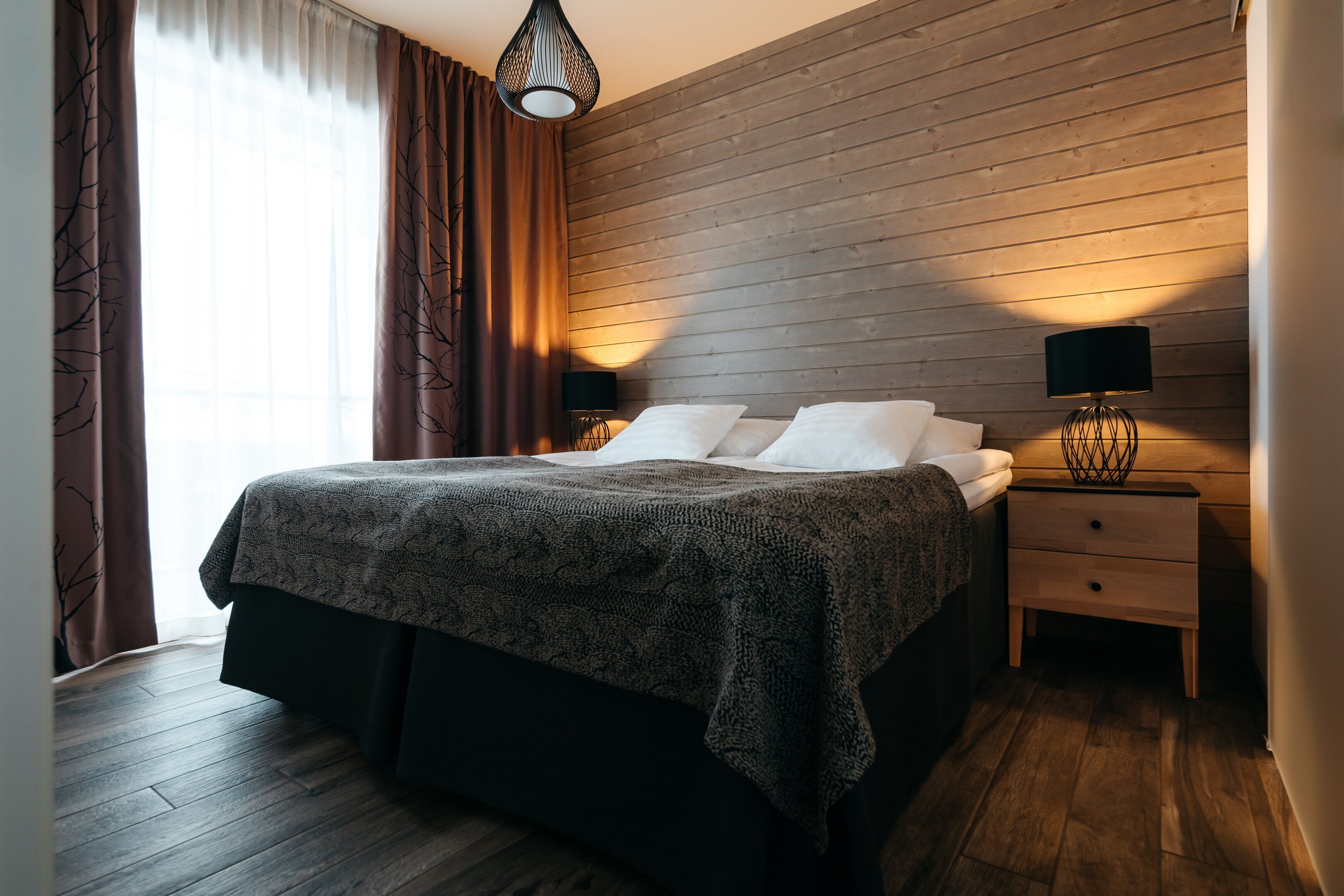 Un dormitorio moderno con una pared de madera, una cama grande con colcha estampada, dos mesitas de noche con lámparas y una ventana con cortinas.