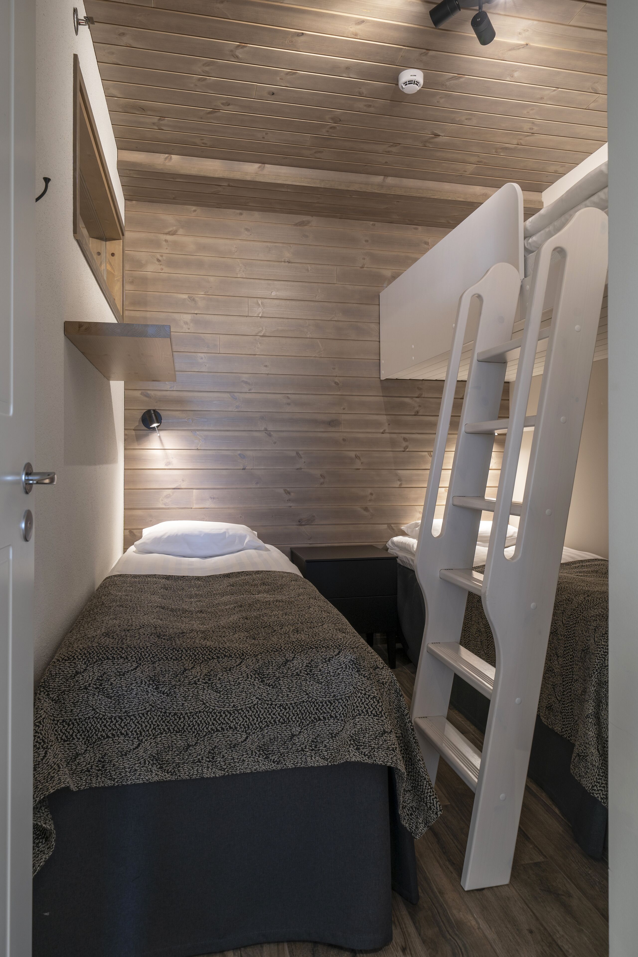 A compact bedroom with a bunk bed, a bedside table, a wall-mounted shelf, and a ceiling light fixture.