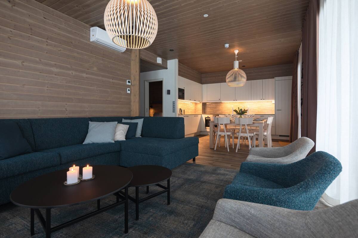 A modern living room with a sectional sofa, a coffee table, and two armchairs. There is a dining area with a table and chairs visible in the background.
