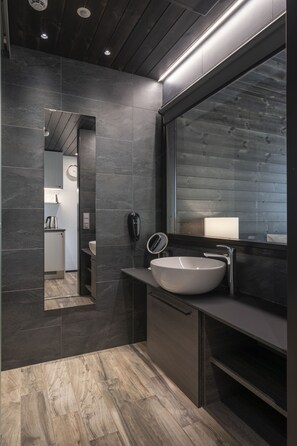 A modern bathroom with dark tiles, a large mirror, a white sink, and a wooden floor.