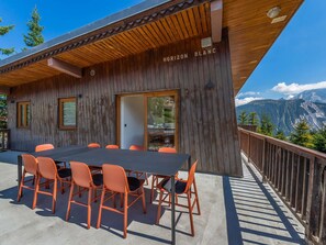 A wooden cabin with a dining area featuring a table and chairs, a view of mountains, and a wooden deck with a railing.