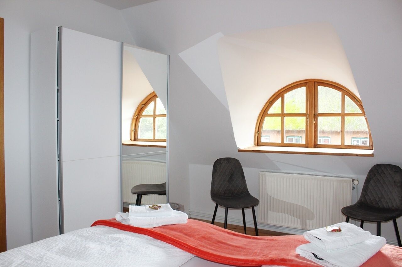 A bedroom with a bed, a large mirror, two chairs, and a window with a view of buildings.