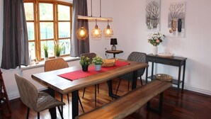 Ein Essbereich mit Holztisch, Stühlen und einer Bank. Es gibt Hängelampen, einen Topfpflanzen und Kunstwerke an der Wand.