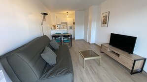 Ett modernt vardagsrum med en grå soffa, ett soffbord i glas, en tv på ett träställ och ett kök i bakgrunden.