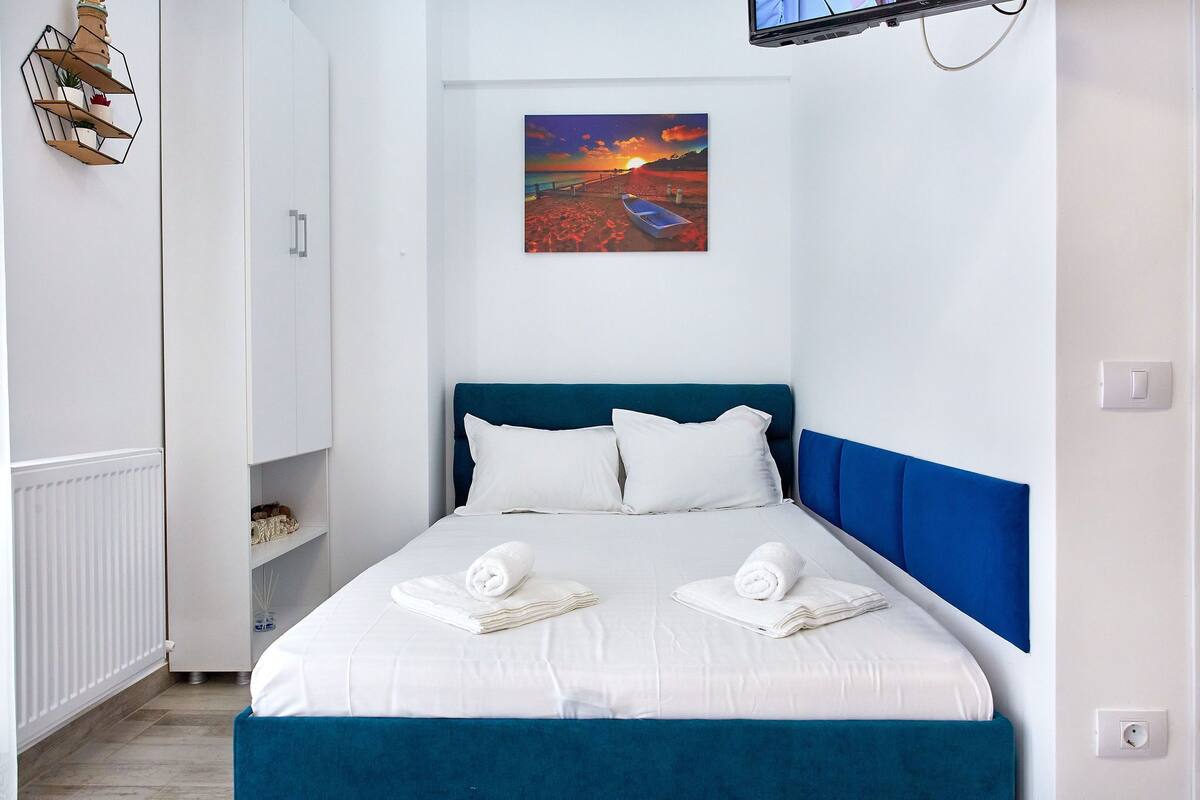 A neatly made bed with white linens and two towels, a blue headboard, and a painting of a sunset on the wall.