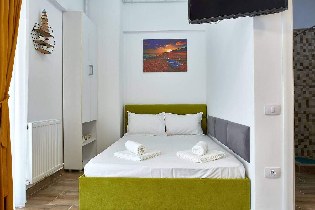 A neatly made bed with white linens and two rolled towels, a green headboard, a painting of a sunset on the wall, and a television mounted above the bed.