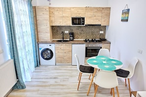 Une cuisine compacte dotée de meubles en bois, d’un four à micro-ondes, d’une machine à café et d’un lave-linge. Un coin repas avec une table et des chaises est également disponible.