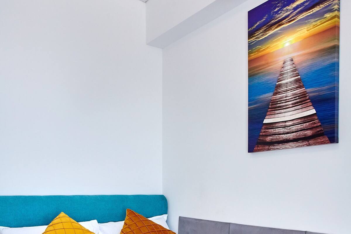 A modern hotel room with a bed, pillows, and a painting of a sunset on the wall.