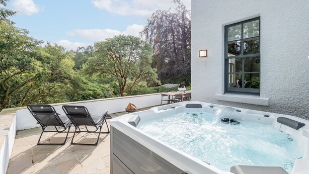 A hot tub with two lounge chairs, a patio table, and a window overlooking a garden.