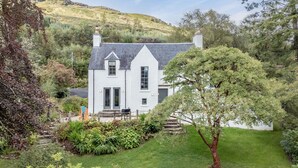A white house with a grey roof, surrounded by lush greenery and a hill in the background.