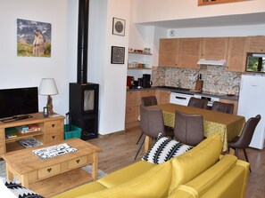 Ein gemütliches Wohnzimmer mit einer Couch, einem Fernseher und einem Kamin. Es gibt einen Essbereich mit Tisch und Stühlen. Die Küche verfügt über Holzschränke und eine geflieste Spritzwand.