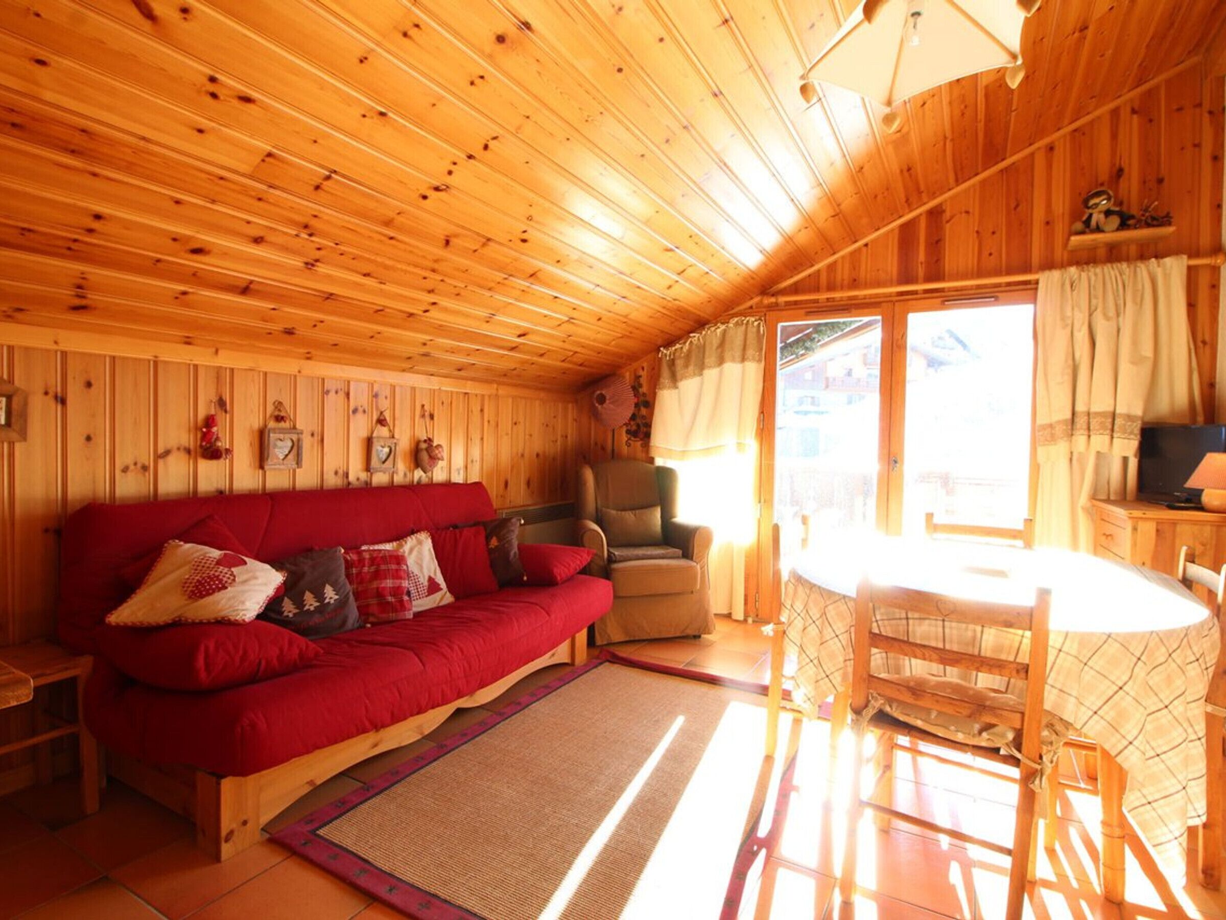 Ein gemütliches Blockhaus-Interieur mit Holzbalkendecke, einem roten Sofa, einem Holzstuhl, einem kleinen Tisch und einem Fenster mit Vorhängen.