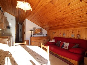 Interior acolhedor de cabana, com teto de madeira, sofá vermelho, cómoda de madeira e área de cozinha.